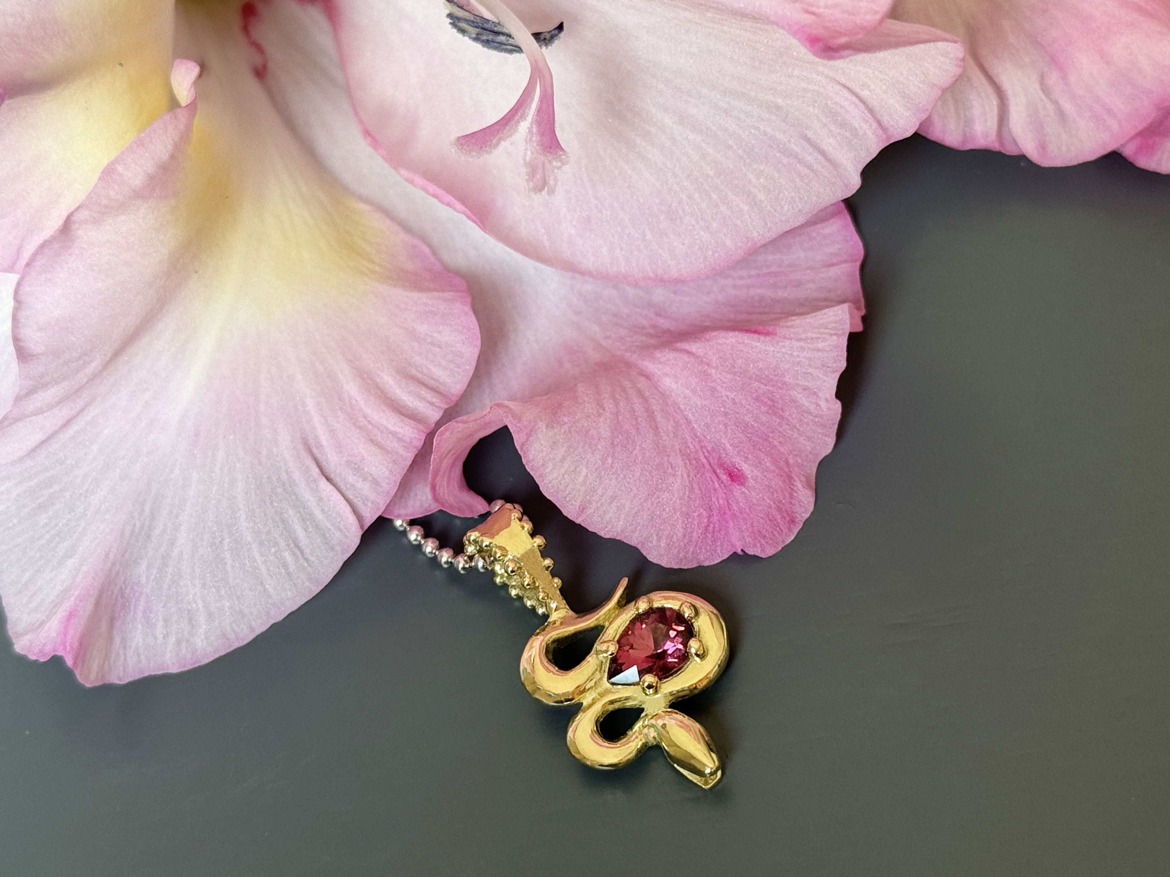 A close-up of the Bohème Serpent Charm in luminous 18 karat yellow Fairmined gold, coiled around a vivid raspberry Rhodolite Garnet. The serpent's intricate details catch the light, set against a dramatic grey background with a soft pink lily accent.