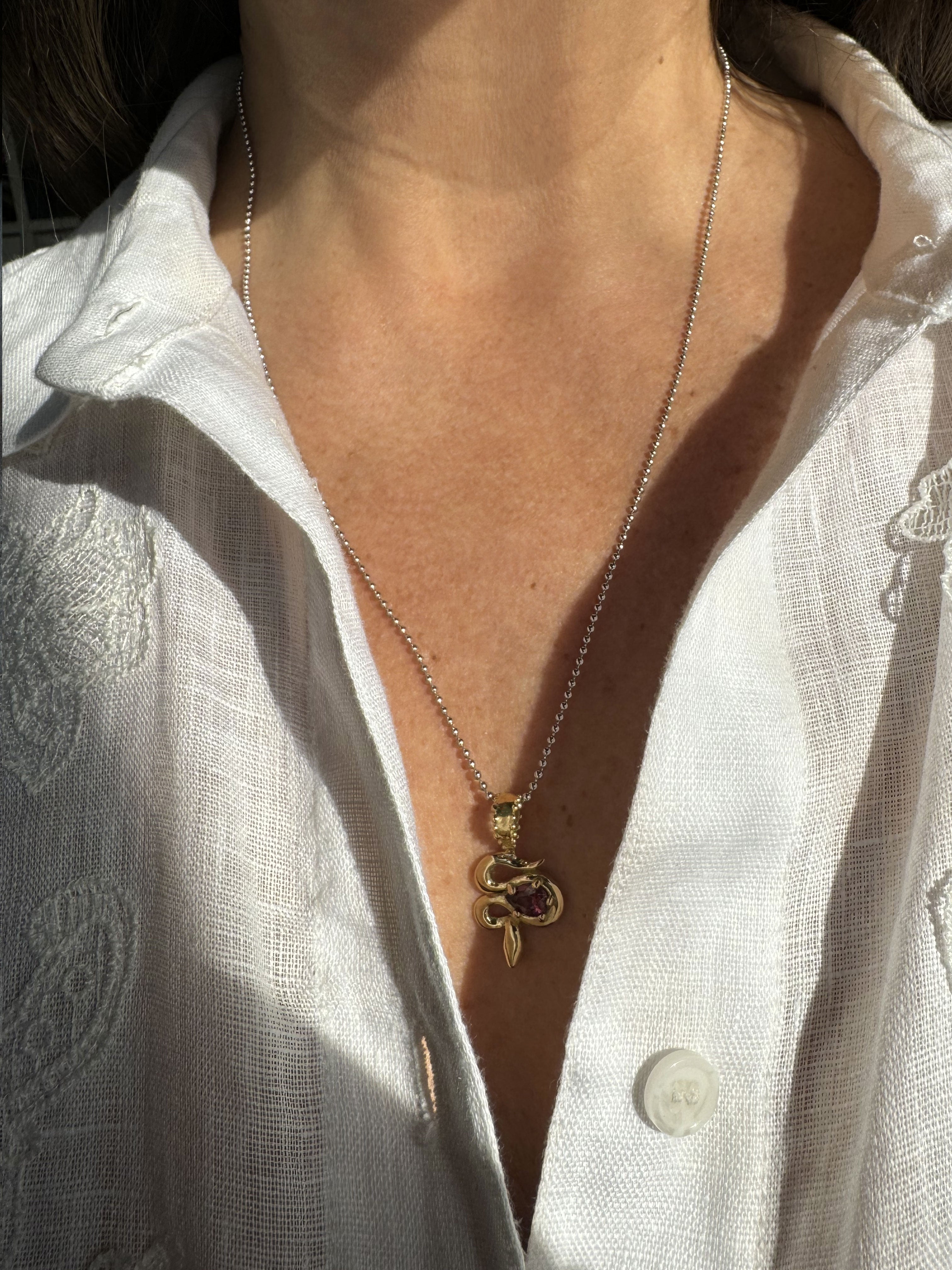 A close-up of the Bohème Serpent Charm in luminous 18 karat yellow Fairmined gold, coiled around a vivid raspberry Rhodolite Garnet. Shown worn, the serpent's intricate details play with the light, nested inside a dramatic deep V of an unbuttoned embroidered white linen shirt.